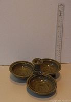 Front view of greenware ceramic spice dish with three small round connected bowls on a raised central pedestal, shown next to a ruler for size reference.