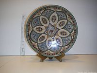 Photo showing entire round ceramic plate with hand-painted traditional colorful design, placed on a stand with a ruler for size reference.