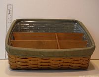 Front angled view of rectangular Longaberger basket with divider sections inside, sage liner on rim, clear protector cover on top, and measuring ruler beside for scale.
