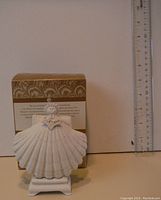 White bisque porcelain Margaret Furlong shell art sculpture with figure and display stand positioned next to ruler and box