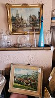 Wide view showing large cityscape painting in ornate gilt frame above smaller village landscape painting on shelf