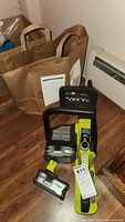 Ryobi chainsaw with two batteries and charger on floor near brown paper bag