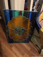 Smaller stained glass panel with large amber flower and green-blue border leaning on floor