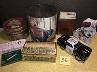 Photo showing the collection of vintage metal tins including large Bulldog football can, Scottish Terrier tin, Police Car toy, and other decorated tins in varied shapes and sizes.