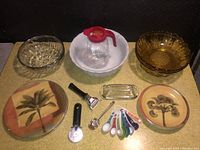 Overview of lot showing vintage glass bowls, ceramic mixing bowl, covered butter dish, juice pitcher, kitchen utensils, decorative plates, measuring cups, and pizza slicer.