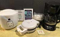Complete lot showing white rice cooker with coffee maker, waffle maker, bag of coffee filters, and two small decorative signs.