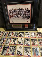 Framed Team Canada gold medal winning team photo from 2002 Olympics shown on black frame with red and white crest pin below photo and plaque. Hockey cards laid out below in rows showing assorted Canada Cup 1976 and Canada Russia Series 1991 cards.