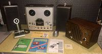 Reel to reel tape recorder with speakers, microphone, tube radio, and manuals as arranged on table