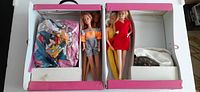 Top-down view of the open pink and white Barbie case with four Barbie dolls arranged in compartments, along with bags of outfits and accessories.