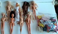 Eight Barbie dolls displayed on white surface, some nude, two in white dresses, and a sealed bag with assorted small doll clothes and accessories.