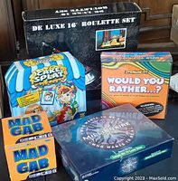 Overview of all games in the lot: De Luxe Roulette Set in black box, Cake Splat, Would You Rather?, Mad Gab, and Who Wants to Be a Millionaire games.