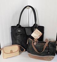 Three bags side by side: a black computer tote bag with new tags behind, a dark green and brown leather handbag in front right, and a small beige woven handbag with brown strap in front left.
