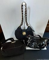 Three handbags displayed together: black quilted with gold medallion by Anne Klein, smooth black fabric by Black House/White Market, shiny patent leather with silver buckles by Fossil
