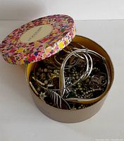 Top-down view of mixed jewelry inside floral patterned round L'Occitane container with lid partially off.