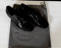 Pair of black leather and suede toe Alexander McQueen Oxford shoes with laces on a white surface, displayed on top of gray dust bag with Alexander McQueen branding.