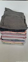Stack of 16 men's button front shirts folded, showing various patterns and colors including stripes, checks and solids with top shirt in faded gray.