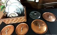 Photo showing multiple pot lids including copper, glass, and stainless steel, along with a woven basket on a black surface.