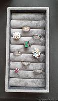 Photo showing 9 sterling silver rings with various stones arranged in a gray ring display tray, stones include pearls, malachite, turquoise, clear rhinestones, and red stones.