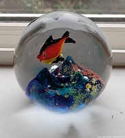 Clear spherical glass paperweight on a window ledge showing a colorful underwater scene with a red and yellow fish above a blue coral-like structure inside.
