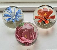 Top-down image showing three spherical glass paperweights with detailed colored flowers inside: orange flower with yellow center, blue flower with bulbous center, and pink flower with multiple petals.