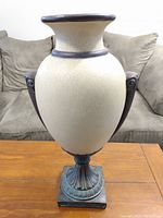 Front view of large cream crackle glaze ceramic vase with dark sculpted handles and ornate square base placed on wooden table.