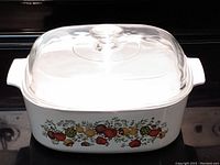 Side view of CorningWare dish showing cultured pattern of colorful fruits and vegetables on white base and clear glass lid with knob handle.
