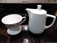 White ceramic Melitta coffee filter holder with handle and ridged interior shown alongside the coffee pot.