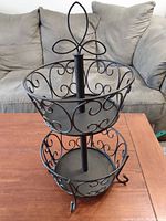 Photo of a black, two-tier metal basket stand on wooden table with sofa background.