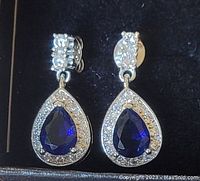 Close-up of sterling silver drop earrings with pear-shaped blue stones surrounded by small white stones, attached to studs with white stones.