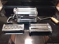 Full view of pasta machine with handle, clamp, and two attachment rollers laid out on a black surface.