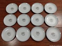Twelve white side plates with identical floral center designs arranged on a table.