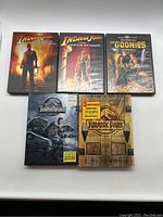 Front view of 6 adventure movie DVDs: Indiana Jones Kingdom of the Crystal Skull (SteelBook), Indiana Jones Temple of Doom, The Goonies, Jurassic World, and Jurassic Park 3-movie collection box set.