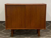 Front elevation of rectangular teak cabinet with two sliding doors closed, inset handles visible, on round tapered legs, positioned on patterned carpet