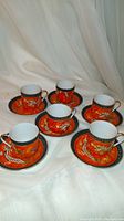 Six orange and black demitasse cups and saucers displayed on white cloth with dragon designs.