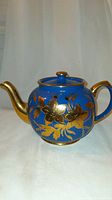 Front view of the blue teapot showing gold floral lily design and gold-spouted curved spout and handle.