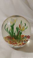 Clear round glass paperweight with visible internal art glass flowers and multicolored pieces at the base, photographed from front.