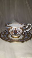 Full view of the Buckingham Palace Diamond Jubilee teacup and saucer set showing robin's egg blue and gold decorative pattern with royal crest on the cup.