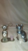 Two chrome cat figurines on a white cloth background. One cat has a clock and a coiled wire behind for holding notes or photos, with a pink pen holder attached to the base. The other cat figurine is sitting with smooth chrome finish and black markings on back.