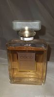 Front view of clear rectangular glass bottle with label Coco Mademoiselle Chanel Paris and frosted glass stopper on top, showing amber liquid inside at close to full level.