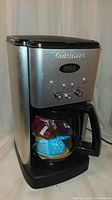 Front angled view of the Cuisinart coffee maker showing the stainless steel body, control panel with timer and switches, and the glass pot filled with packaging inside.
