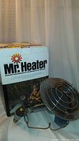 Photo showing the Mr. Heater portable radiant heater head with the 8-inch round heating element and propane connection next to the original branded box.