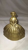 Front view of Southern Belle brass bell showing seated female figure in wide dress with floral patterns and holding a basket.