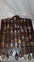 Full view of the dark wood plaque with 24 souvenir teaspoons hung in three rows. Spoons feature various tops and are evenly spaced.