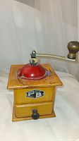 Vintage wooden spice or coffee grinder with red metal top, metal crank handle with wooden knob, metal drawer in front, and vintage label on front.