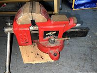 Side view of red Little Daddy 3-1/2" bench vise mounted on a wooden board, showing the jaws and handle.