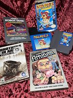 Overview of game cartridges and boxes: Nintendo NES Puss'n Boots, Atari Amidar and Robot Tank, Brothers in Arms PC box seen.