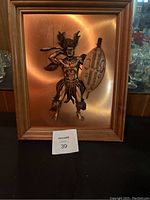 Photo of a framed 3D metal artwork depicting a tribal warrior holding a spear and shield on a copper-toned background. The wooden frame and details of metalwork are visible.