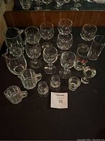 49 pieces of mixed glassware displayed on black surface including various sizes and types of shot glasses, measuring glasses, drink glasses, and wine glasses with some etched floral patterns.