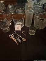 Photo of mixed bar accessories including two wine glasses, two ice buckets, a cut glass decanter, shot glasses, bottle openers, corkscrew, spoon, and other tools on a black surface.