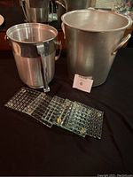Two metal ice buckets, one larger and one smaller, placed side by side on a black surface. Five glass coasters with textured underside arranged in front, showing clear glass and small feet. The smaller bucket has a tape piece on the side possibly holding a tong.